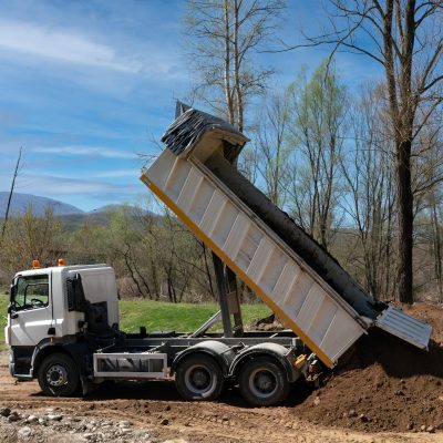 White dump truck unloading soil at a construction site. Heavy machinery performing earth works, backfilling for a landscape architecture project. Industrial earthmoving operation.